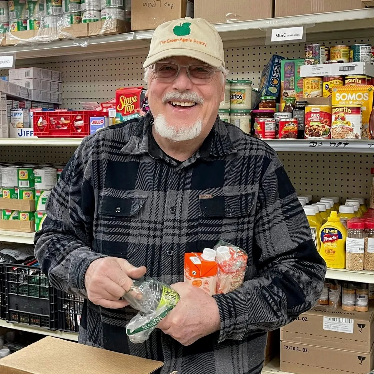 Friendly volunteer in a Green Apple Pantry hat smiling among the pantry shelves